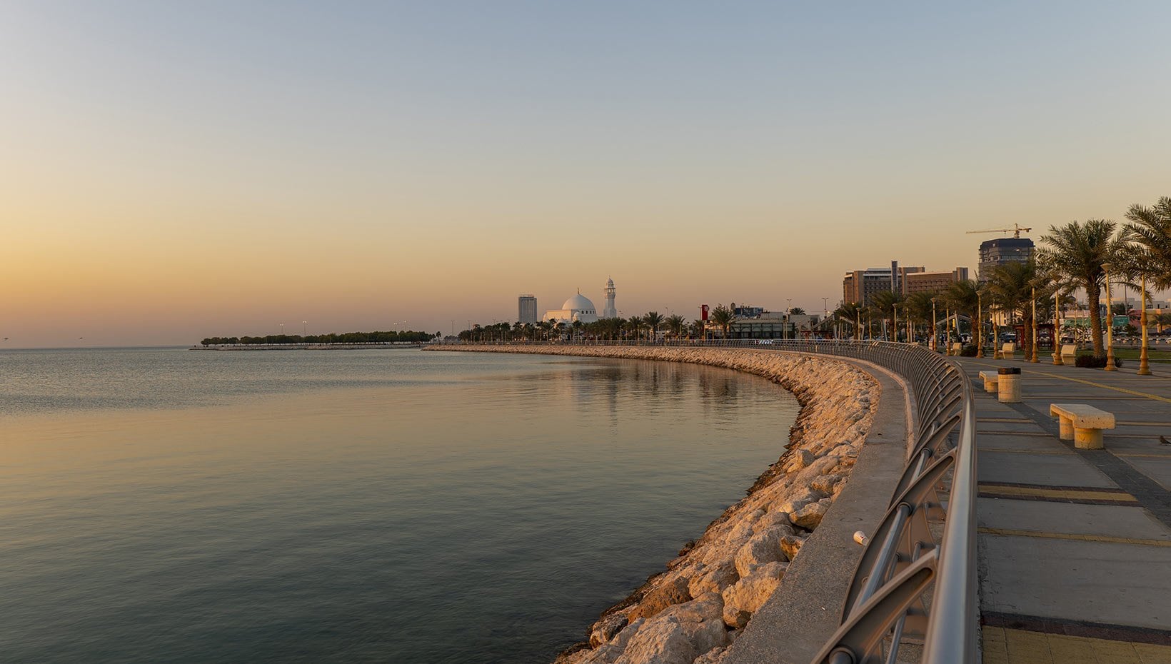 Scenic waterfront promenade at sunset with palm trees and cityscape | MSC Cruises Scenic waterfront promenade at sunset with palm trees and cityscape | MSC Cruises