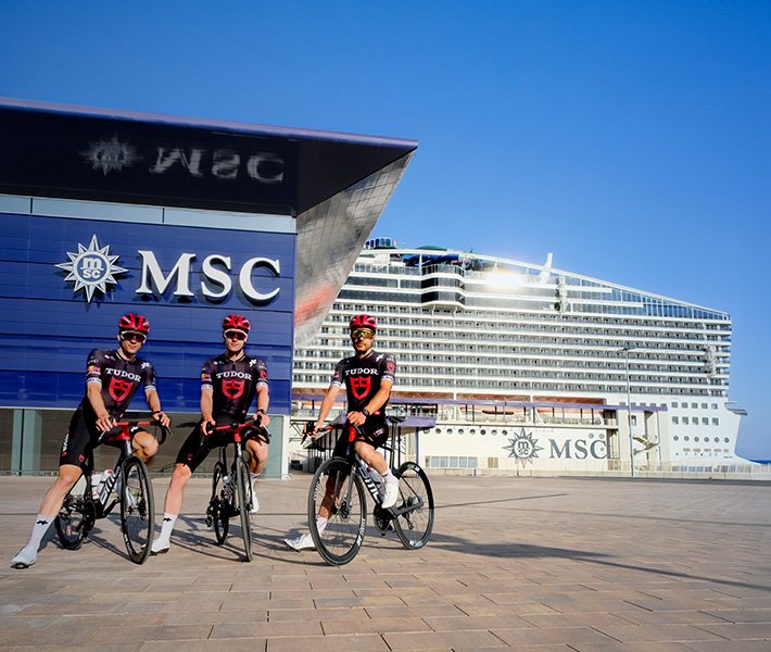 Tres ciclistas con indumentaria de marca frente a una terminal de cruceros de MSC y un barco, bajo un cielo azul despejado | MSC Cruceros