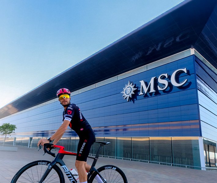 Un ciclista con indumentaria de MSC frente a un edificio moderno de MSC | MSC Cruceros Un ciclista con indumentaria de MSC frente a un edificio moderno de MSC | MSC Cruceros